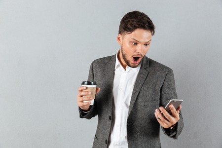 Portrait of a shocked businessman looking at mobile phone while holding cup of coffee to go isolated over gray backgroundの写真素材