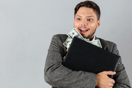 Portrait of a happy businessman hugging briefcase full of money banknotes isolated over gray backgroundの写真素材