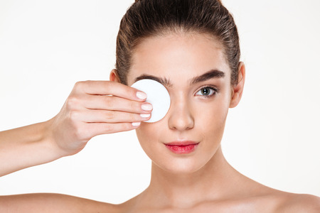 Close up photo of brunette beautiful woman removing her eye makeup with lotion and cotton pad, isolated over white backgroundの写真素材