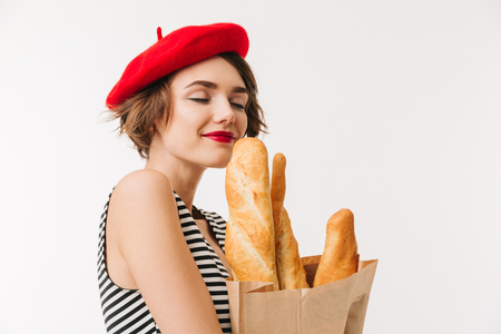 Portrait of a pretty woman wearing beret smelling paper bag with long loaf isolated over white backgroundの写真素材