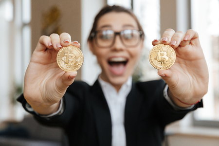 Image of young excited business lady showing her golden bitcoins sitting indoors coworking. Looking camera. Focus on bitcoins.の写真素材