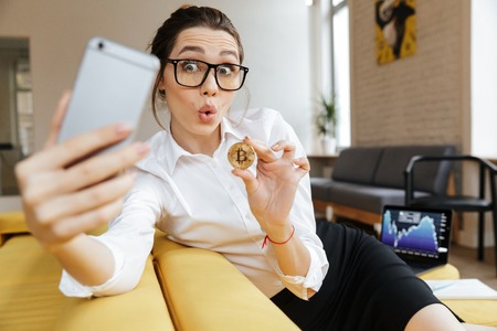 Photo of young excited emotional business lady talking by mobile phone sitting indoors coworking make selfie showing her golden bitcoin. Looking aside.の写真素材