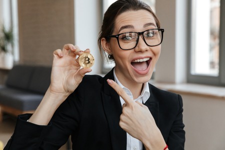Image of young excited business woman showing her golden bitcoin sitting indoors coworking. Looking camera pointing.の写真素材