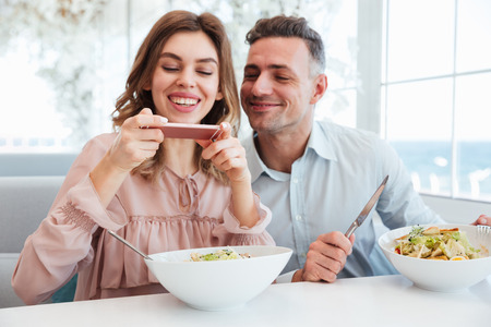 Photo of cheerful married couple having dinner and woman photographing her meal on smartphone before eatingの写真素材