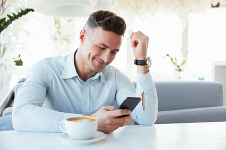 Image of happy elegant man sitting alone in city cafe with cup of cappuccino and typing text message using black mobile phoneの写真素材