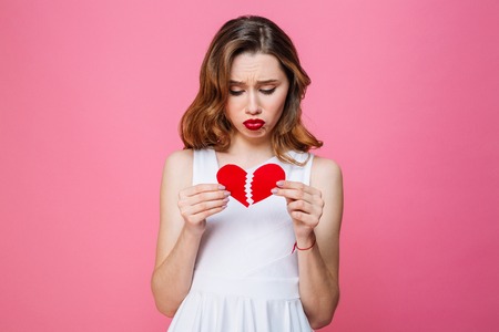 Photo of young sad woman standing isolated over pink background holding broken heart. Looking aside.の写真素材
