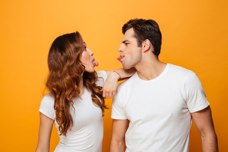 Portrait of funny man and woman in white t-shirts smiling and looking on each other with tongues sticking out isolated over yellow backgroundの写真素材