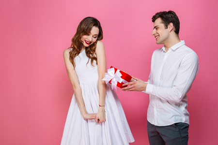 Portrait of a happy smartly dressed couple holding present box isolated over pink background, man giving present to his girlfriendの写真素材