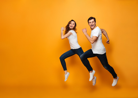 Young lovely cheerful couple posing together on camera while running or jumping along yellow...