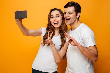 Young lovely friends smiling and making selfie on smartphone while showing victory signs together, over yellow backgroundの写真素材