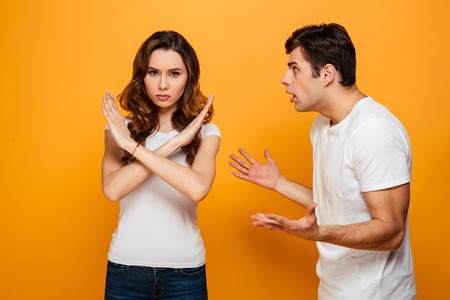 Displeased man screaming on his girlfriend which showing stop gesture and looking at the camera over yellow backgroundの写真素材
