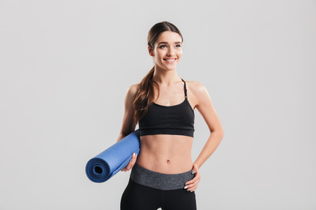 Attractive young sportswoman holding yoga mat and looking on camera isolated over gray backgroundの写真素材