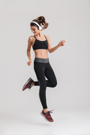 Full-length image of strong-willed female fitness trainer of instructor in sportswear jumping with wireless headphones isolated over gray backgroundの写真素材