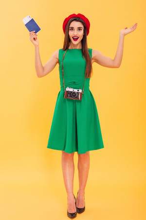 Photo of excited young woman photographer tourist standing isolated over yellow background holding camera and passport with tickets.の写真素材