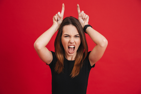 Portrait of crazy woman 20s wearing black screaming and playfully showing horns with two fingers isolated over red backgroundの写真素材
