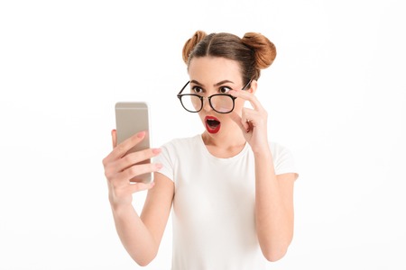 Portrait of an astonished casual girl in eyeglasses looking at mobile phone isolated over white backgroundの写真素材