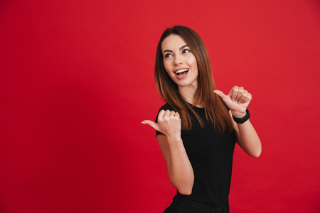 Smiling woman 20s with long brown hair posing at camera and pointing fingers backward on copyspace isolated over red backgroundの写真素材