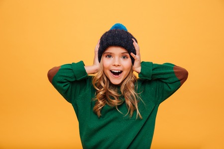 Surprised happy Young girl in sweater and hat holding her head while looking at the camera over orange backgroundの写真素材