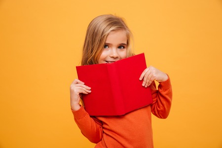 Smiling young girl in sweater hiding behind the book and looking at the camera over orange backgroundの写真素材