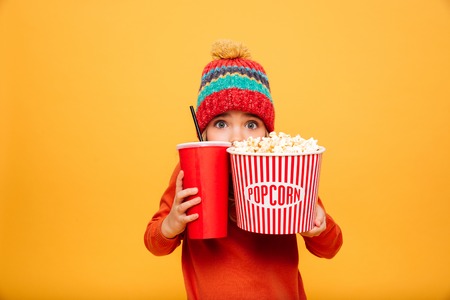 Scared Young girl in sweater and hat hiding behind the popcorn and plastic cup while looking at the camera over orange backgroundの写真素材