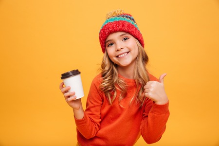 Smiling Young girl in sweater and hat holding cup of tea and showing thumb up while looking at the camera over orange backgroundの写真素材