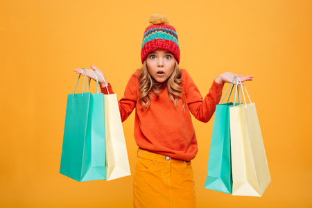 Confused Young girl in sweater and hat holding packages while looking at the camera over orange backgroundの写真素材