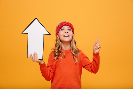 Happy Young girl in sweater and hat holding paper arrow while pointing and looking up over orange backgroundの写真素材