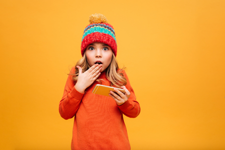 Shocked Young girl in sweater and hat holding smartphone while covering her mouth and looking at the camera over orange backgroundの写真素材