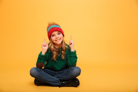 Happy Young girl in sweater and hat sitting on the floor while pointing up and looking at the camera over orange backgroundの写真素材