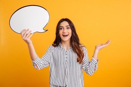 Surprised happy brunette woman in shirt holding blank speech bubble and looking at the camera over...