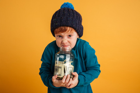 Image of angry displeased little redhead boy with freckles standing isolated over yellow background wearing warm hat. Looking camera holding jar with money.の写真素材
