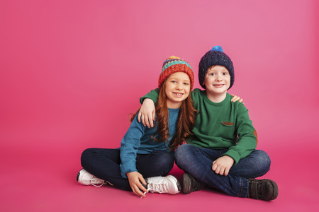 Cheerful brother and sister in knitted clothes hugging and looking camera isolated over pink...