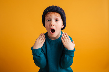 Photo of shocked little redhead boy with freckles standing isolated over yellow background wearing warm hat. Looking camera.の写真素材