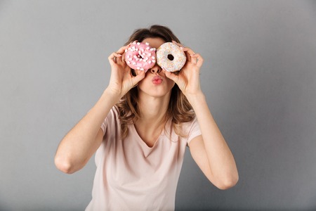 Playful woman in t-shirt having fun while holding donuts on her eyes and sends air kiss over grey backgroundの写真素材