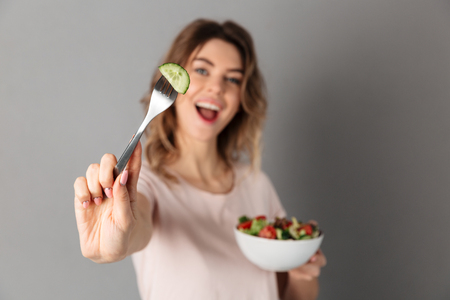 Playful woman in t-shirt holding plate with fresh vegetables and showing cucumber at camera. Focus on fork with cucumberの写真素材