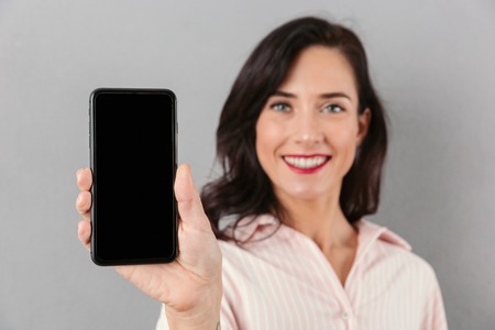 Close up of a smiling businesswoman showing blank screen mobile phone isolated over gray backgroundの写真素材