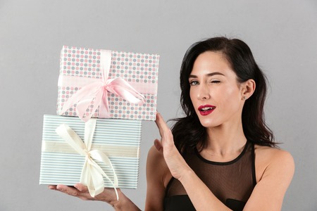 Portrait of an attractive woman dressed holding stack of present boxes and winking isolated over gray backgroundの写真素材