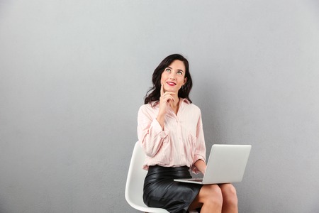 Portrait of a pensive businesswoman looking away at copy space while sitting in a chair and using laptop computer isolated over gray backgroundの写真素材
