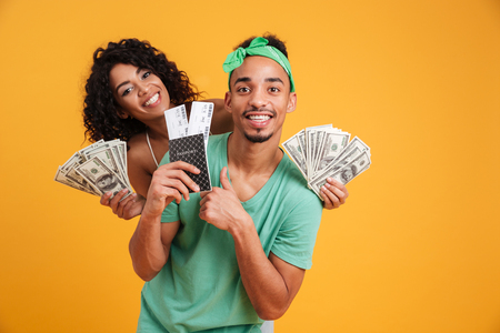 Portrait of a happy young african couple showing passport with flying tickets while holding bunch of money banknotes isolated over yellow backgroundの写真素材