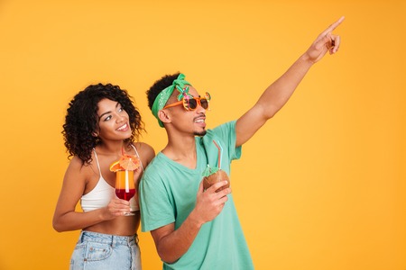 Portrait of a smiling young african couple dressed in summer clothes drinking cocktails while pointing finger away isolated over yellow backgroundの写真素材
