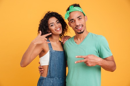 Portrait of a happy young african couple dressed in summer clothes hugging and showing peace gesture isolated over yellow backgroundの写真素材