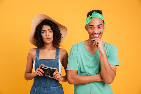 Portrait of a confused young african couple holding photo camera isolated over yellow backgroundの写真素材