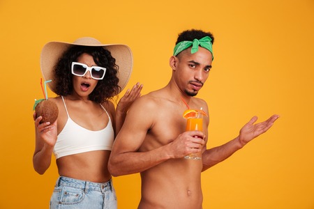Portrait of a confused young african couple in summer clothes holding cocktails and looking at camera isolated over yellow backgroundの写真素材