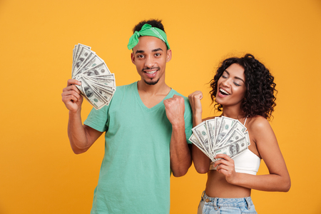 Portrait of a satisfied young african couple in summer clothes showing bunch of money banknotes isolated over yellow backgroundの写真素材
