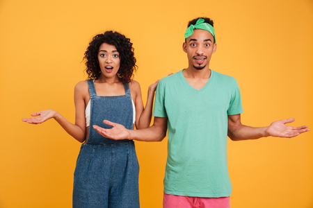 Portrait of a confused young african couple dressed in summer clothes shrugging shoulders isolated over yellow backgroundの写真素材