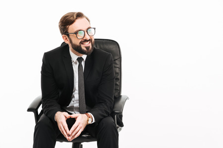 Portrait of happy office worker in suit and eyeglasses smiling while sitting on black armchair and looking aside on copyspace isolated over white wallの写真素材