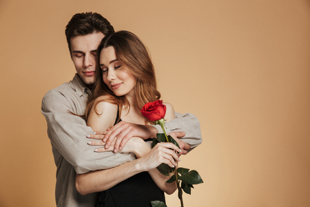 Portrait of a happy loving couple hugging while holding red rose isolated over beige backgroundの写真素材