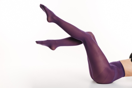 Side view cropped image of a slim young woman dressed in lingerie and violet tights posing while laying with legs raised isolated over white backgroundの写真素材