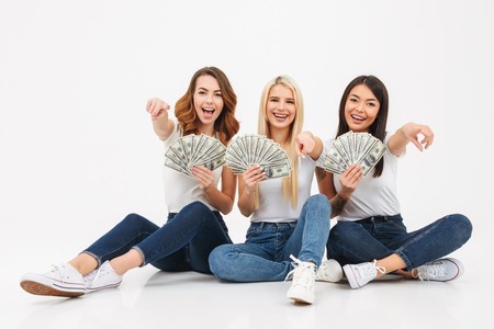 Image of three young cute pretty girls friends sitting isolated over white background looking camera showing money pointing.の写真素材