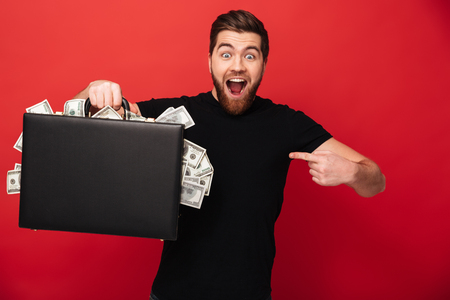 Image of handsome bearded man standing isolated over red background wall holding suitcase full of money pointing. Looking camera.の写真素材